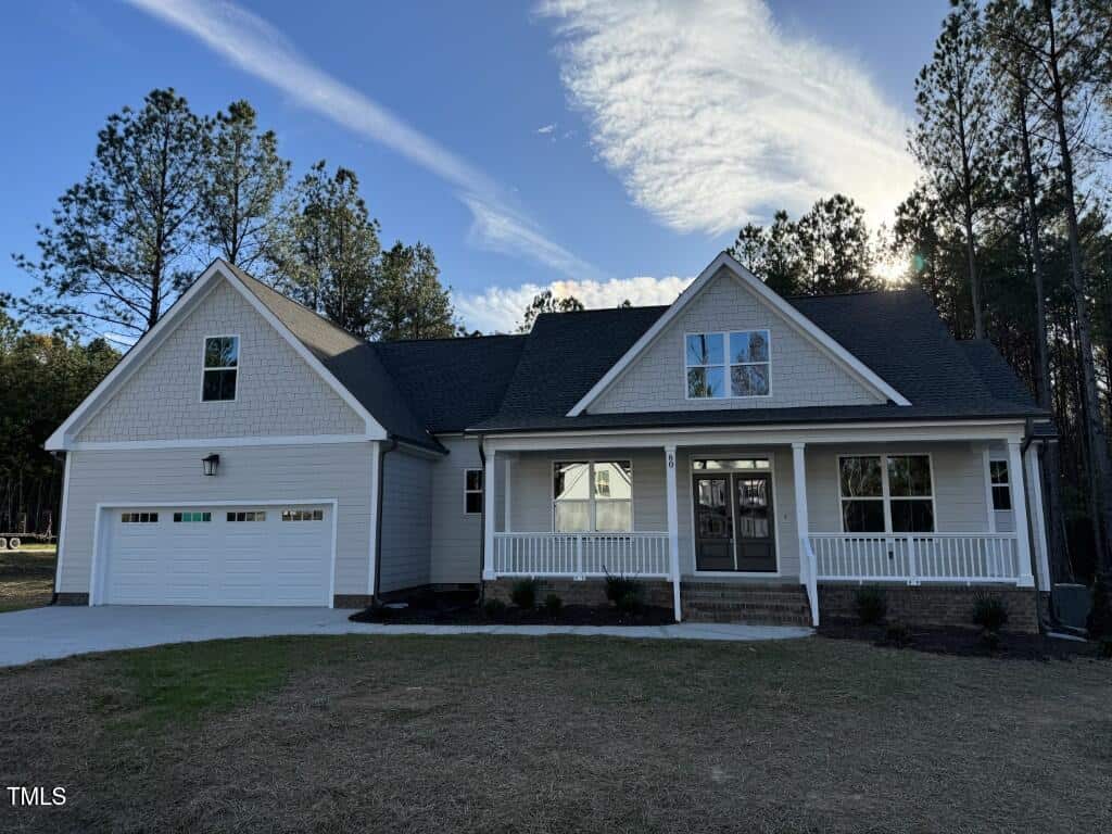 A modern single-story house with gray siding, a covered front porch, double front doors, and a two-car garage, set against a backdrop of tall pine trees under a partly cloudy sky.