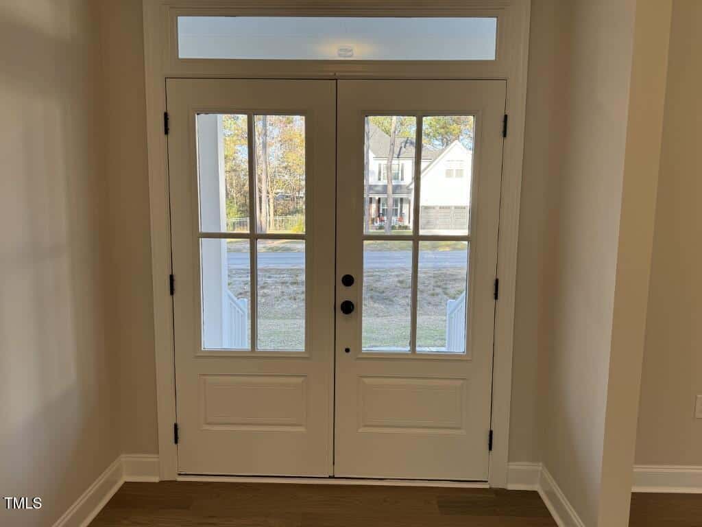 A set of white double doors with glass panes opens to a view of a street and houses across the road, with trees and grass visible outside. Sunlight streams in through the glass panels and transom window above.