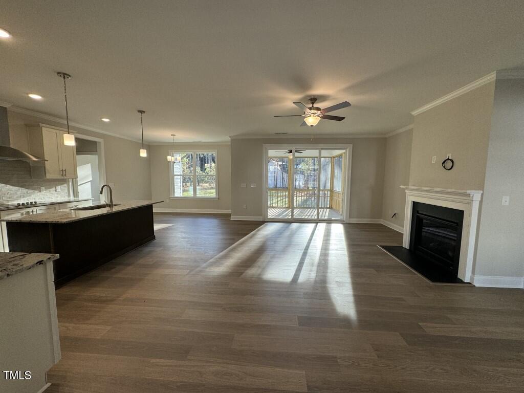 Open-concept living space with wood flooring, kitchen island and pendant lights on the left, dining area with windows in the center, fireplace on the right, ceiling fan, and glass doors leading to a screened porch.