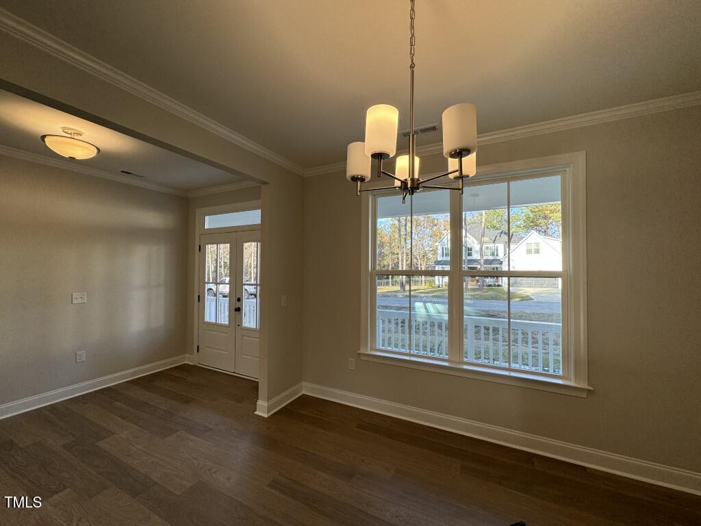 A bright, empty room with wood floors, beige walls, a large window facing outside, a modern chandelier, and a white front door with glass panels. Natural light fills the space, and houses are visible through the window.