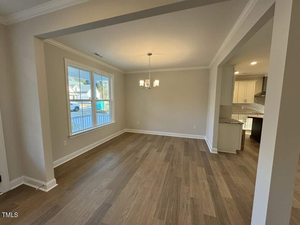 A spacious, empty dining room with light beige walls, wood flooring, a large window, and a modern chandelier. The room opens to a kitchen with white cabinets and stainless steel appliances.