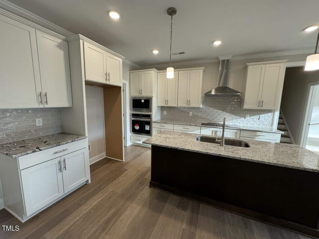 Modern kitchen with white cabinets, stainless steel appliances, a center island with a sink, granite countertops, tile backsplash, wood flooring, and recessed ceiling lights. Stairs are visible in the background.