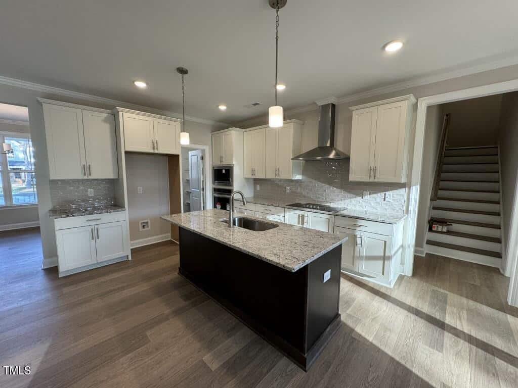 Modern kitchen with white cabinets, stainless steel appliances, a central island with sink, pendant lights, light gray backsplash, wood flooring, and a staircase visible at the right side. Natural light enters from the left.