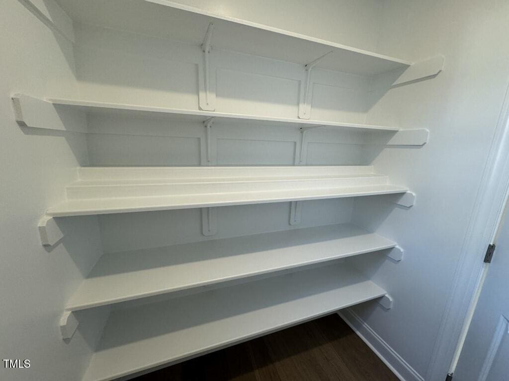 Four empty white shelves are mounted on a white wall in a small pantry or storage room with a dark wood floor and a white door on the right.
