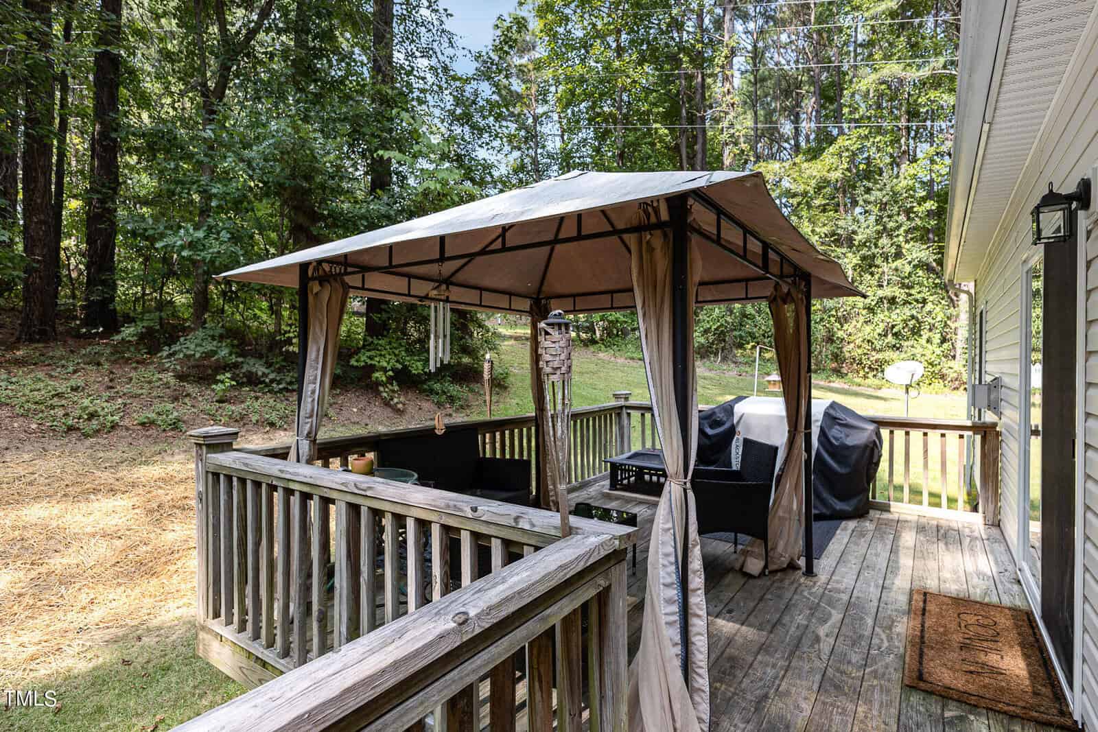 A wooden deck with a gazebo featuring a beige canopy and draped sides. Inside, there are black chairs and a small table. Nearby is a grill, and the deck is surrounded by trees and greenery in the background.