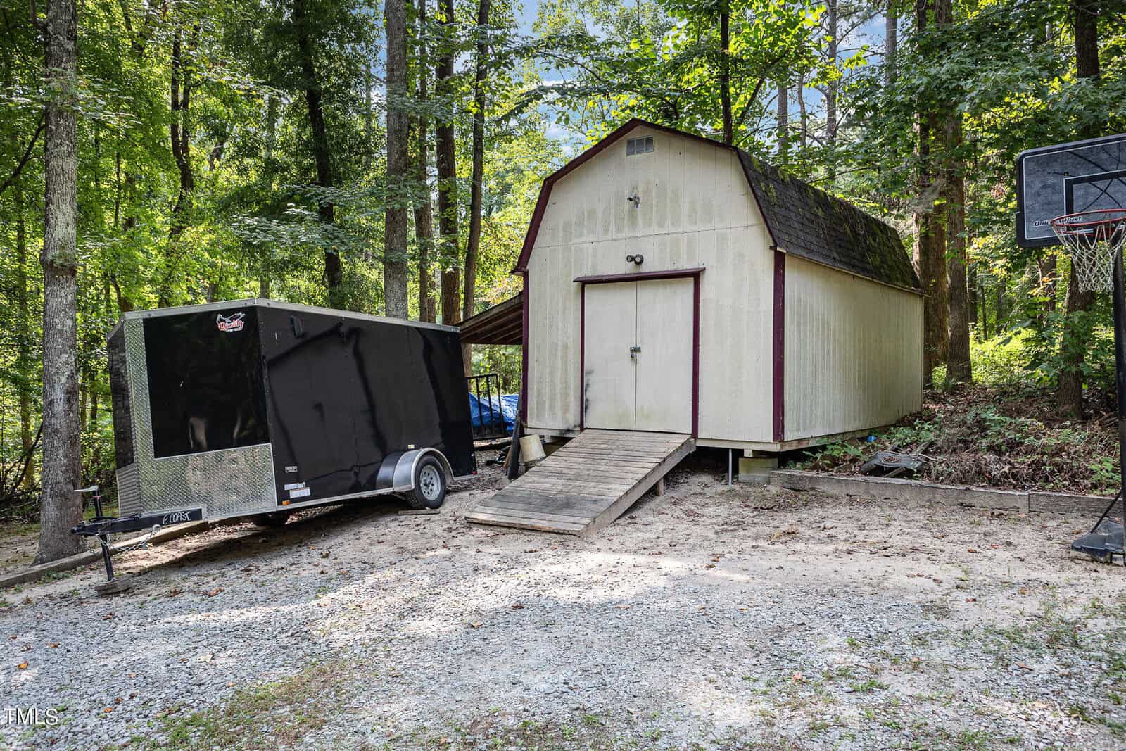 A gravel driveway leads to a beige shed with a brown roof, surrounded by trees. A black trailer is parked beside the shed, and a basketball hoop stands nearby. A wooden ramp provides access to the shed's double doors.