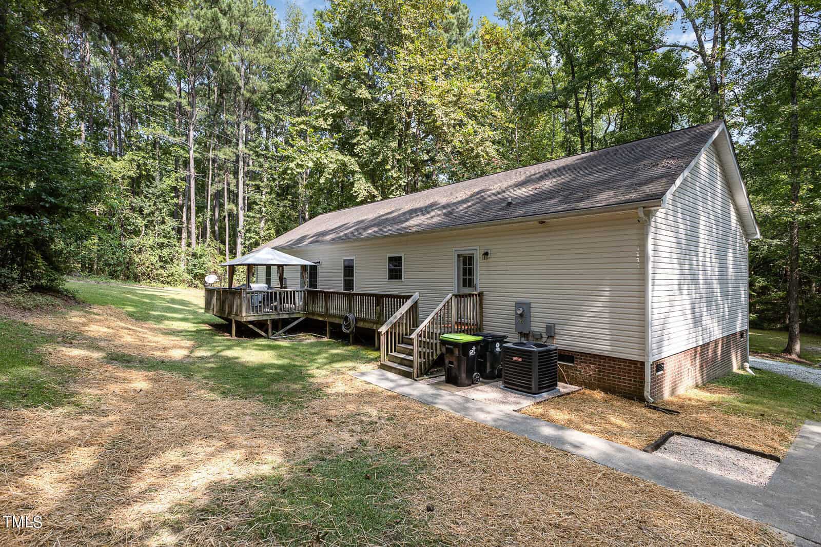 A single-story house with white siding, located beside a grassy yard and surrounded by tall trees. A wooden deck extends from the house with patio furniture. Several garbage bins are placed near the house. A narrow driveway is visible to the right.