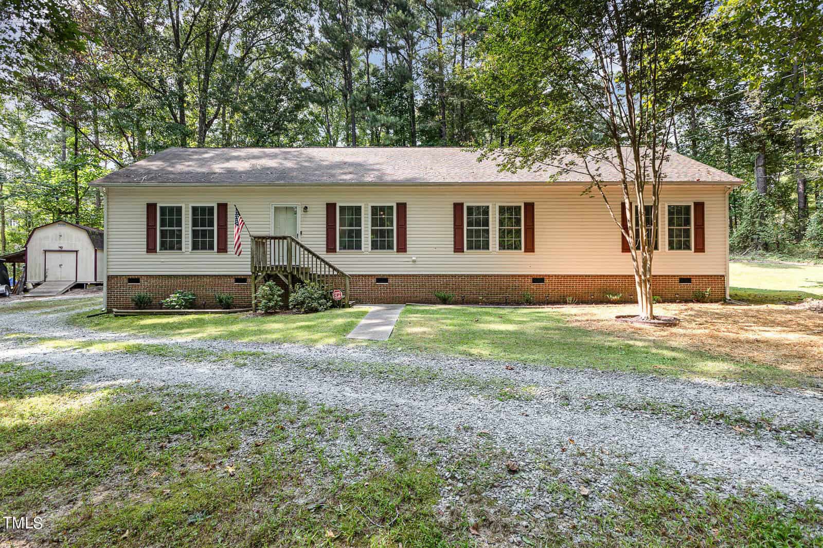 A single-story house with beige siding and dark brown shutters. A small front porch with stairs leads to the entrance. A gravel driveway runs in the foreground. Trees and a red storage shed are visible in the background.