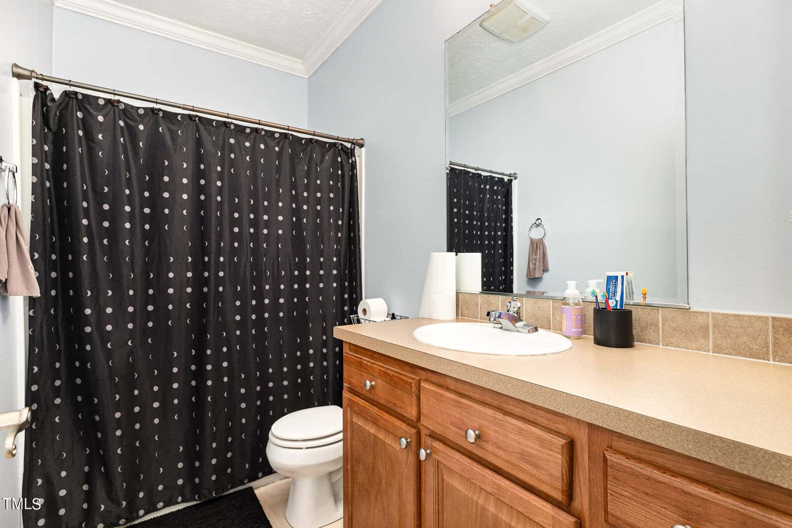A bathroom with a sink, wooden cabinet, and a countertop holding toiletries. A toilet is beside it. A mirror hangs above the sink, and a shower with a black polka dot curtain is in the background. The walls are light blue.