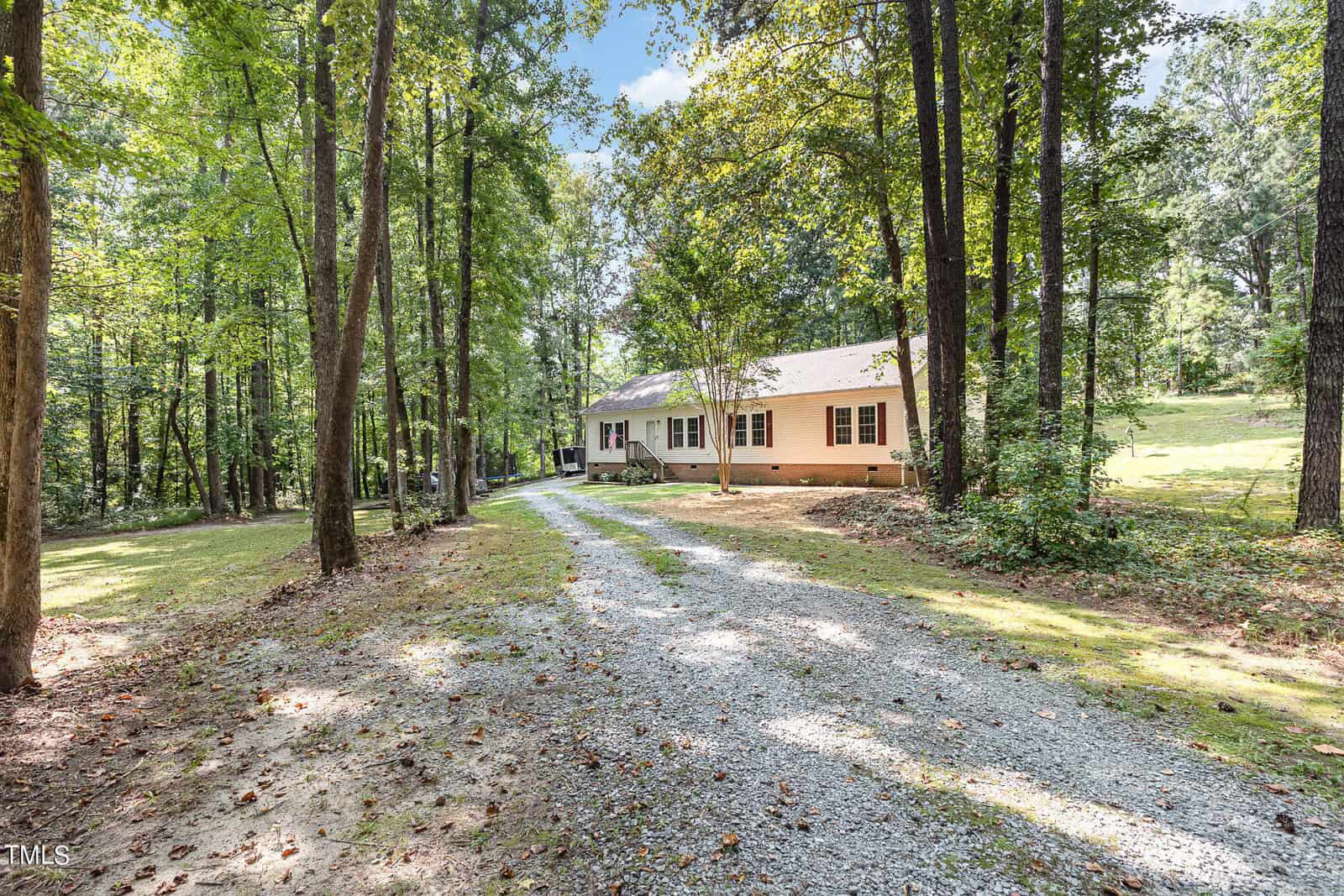 Nestled in a wooded area, this single-story house at 104 Dove Road in Creedmoor features light siding and dark shutters. A gravel driveway winds through tall trees and a grassy yard, leading to the charming home under a clear, blue sky.