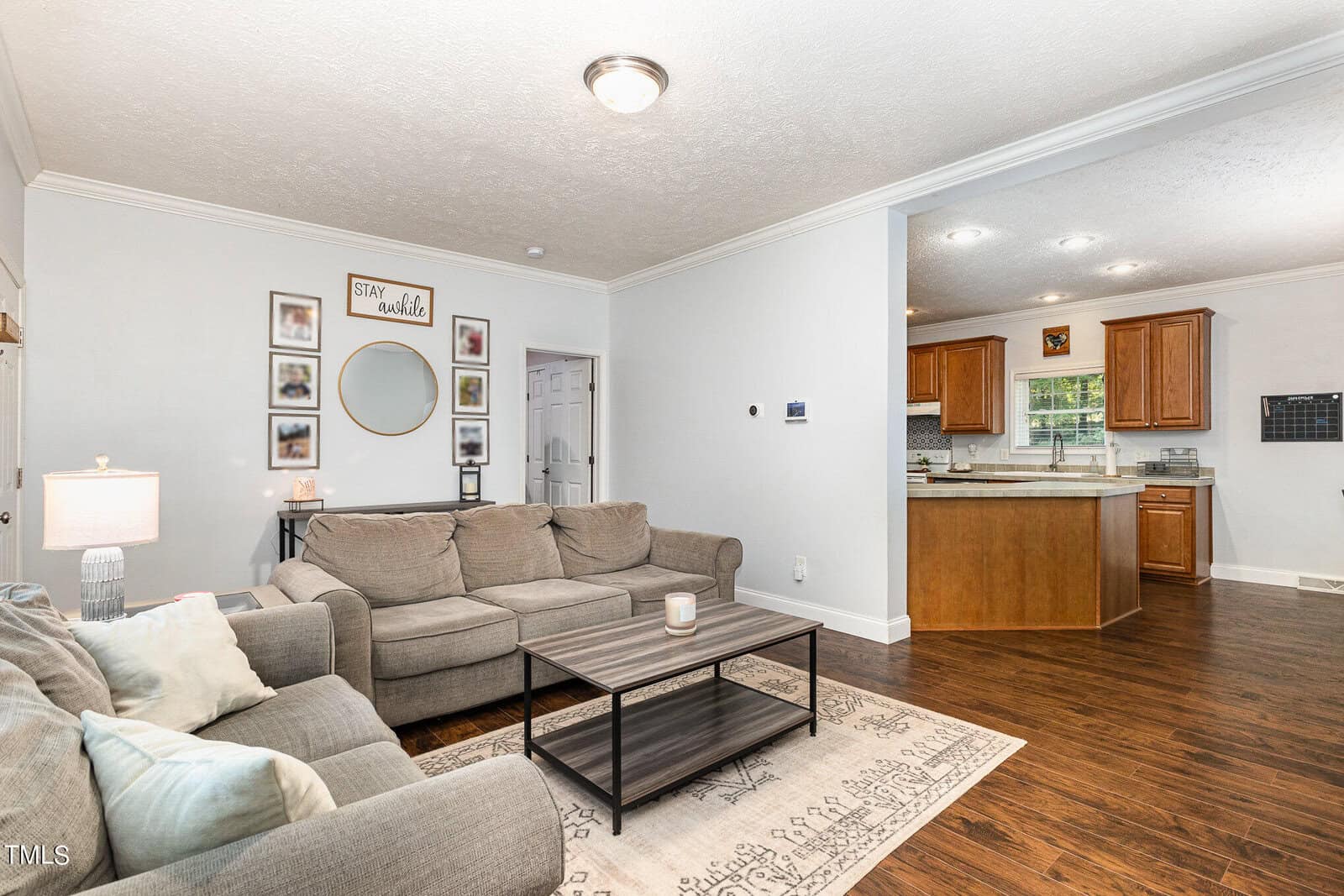A spacious living room with a gray sectional sofa, wooden coffee table, and decorative wall art. A kitchen with wooden cabinets is visible in the background. The room features hardwood floors and neutral-colored walls.