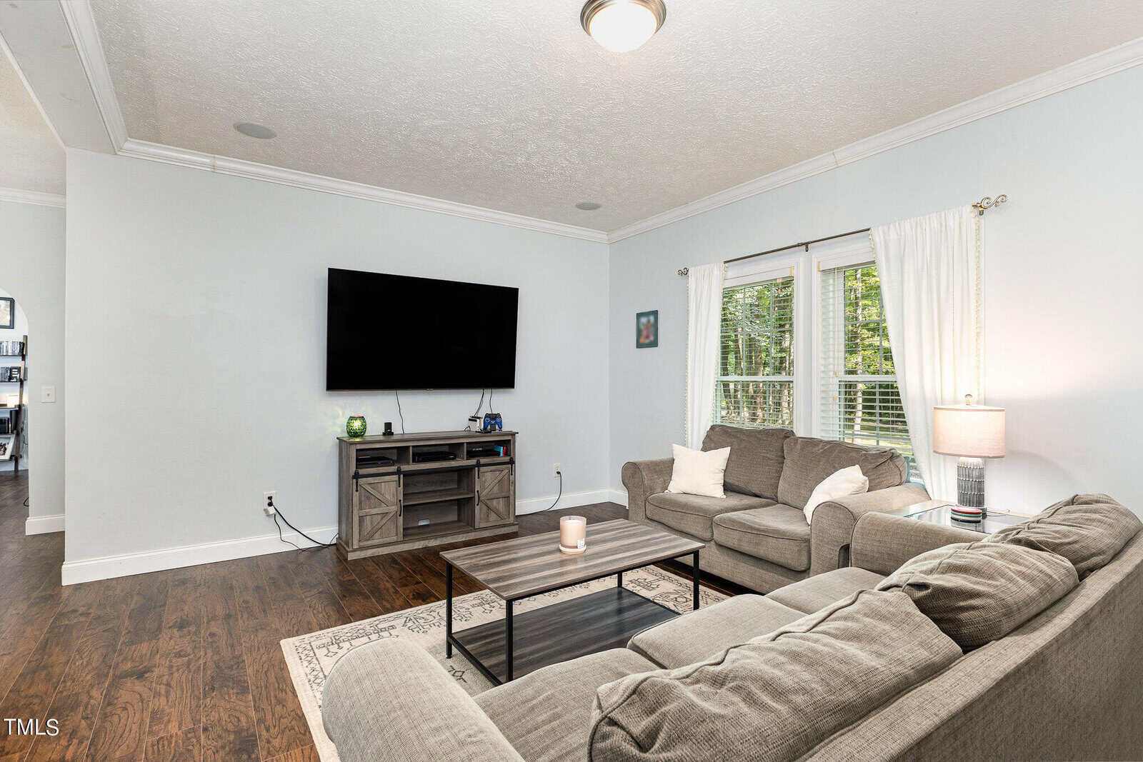 A living room with a gray L-shaped sofa, a wooden coffee table, and a flat-screen TV mounted on the wall. The room has light-colored walls, hardwood floors, a window with white curtains, and a patterned area rug.