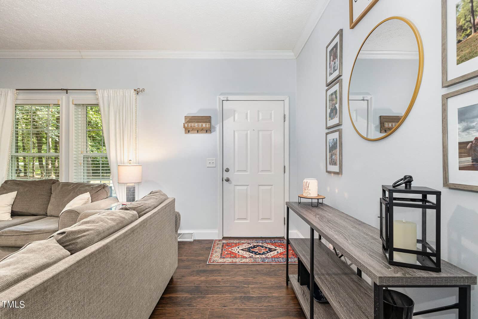 A living room with a gray sofa, a wooden floor, and a rectangular wooden table. The wall has a round mirror and framed photos. A door is visible in the background with a small rug in front of it. There's a window with white curtains to the left.