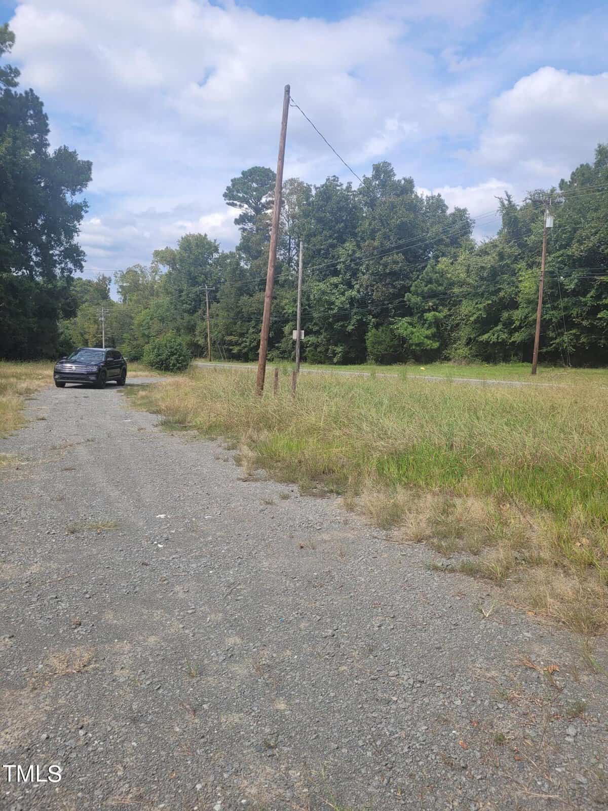 A black car is parked on a gravel path next to a grassy area. Utility poles with wires run alongside the road. The background features trees and a partly cloudy sky.