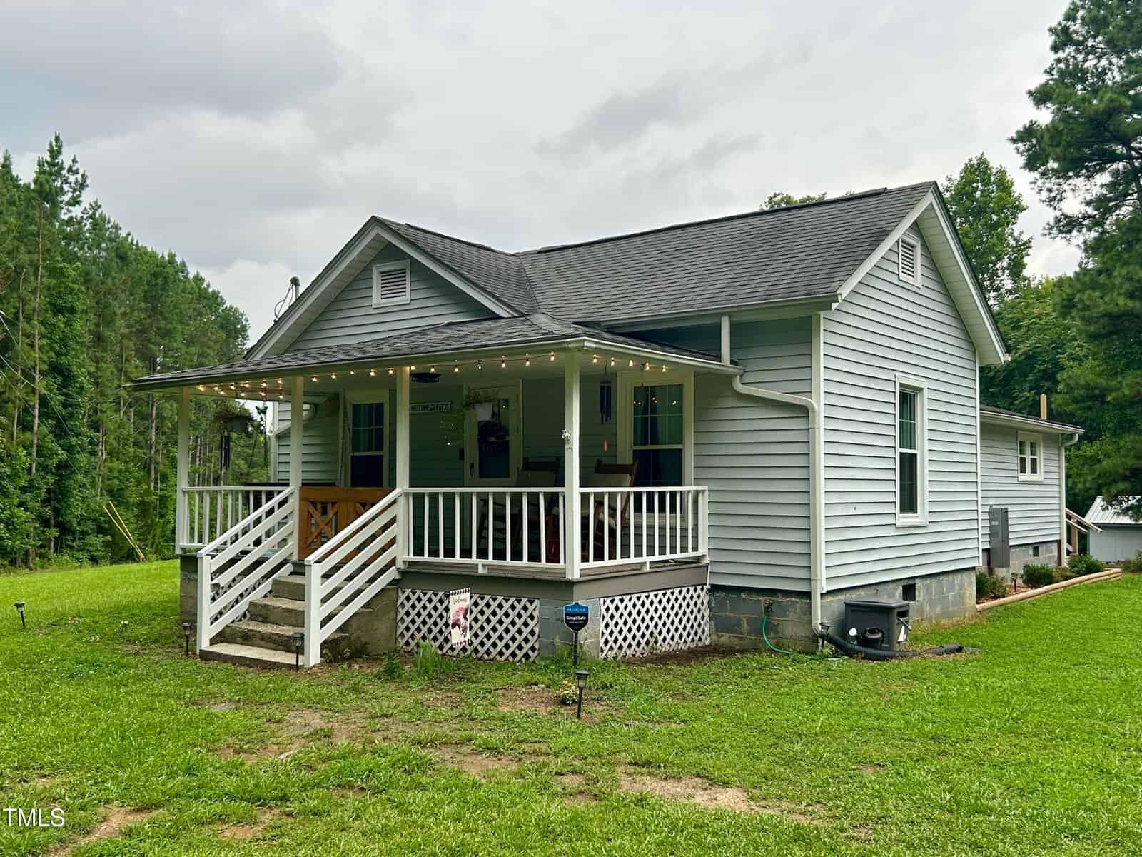 A small, light gray house with a dark roof is pictured. It has a covered front porch with string lights, white railing, and steps leading to the lawn. The house is surrounded by green grass and trees, under a cloudy sky.