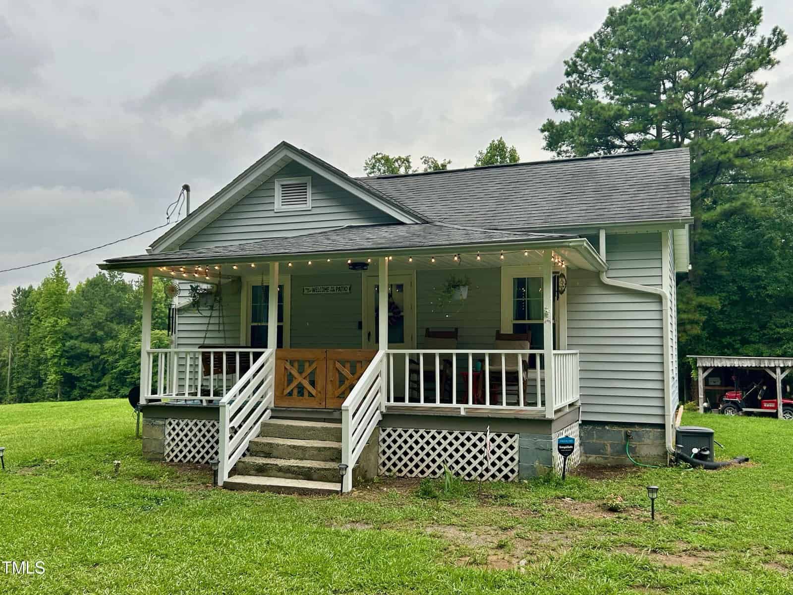 A small, single-story house with a porch, featuring a wooden railing and steps leading up to the entrance. The yard is grassy, surrounded by trees, and a carport with two vehicles is visible on the right. The sky is cloudy.