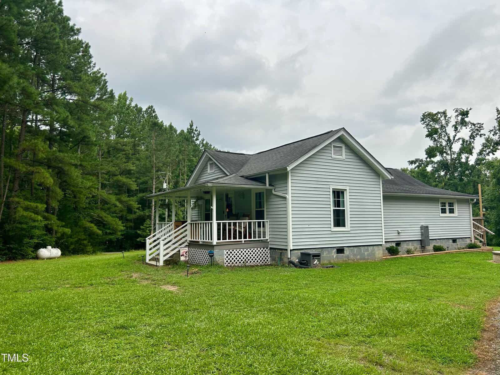 A small, single-story gray house with a dark roof is surrounded by lush green grass and trees. It has a covered porch with white railings. The sky is overcast.