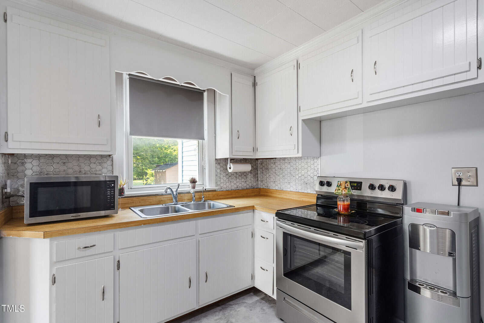 A small kitchen with white cabinets, a stainless steel stove, and a microwave. There's a double sink under a window with a gray blind. A gray backsplash and a compact water dispenser are also visible.