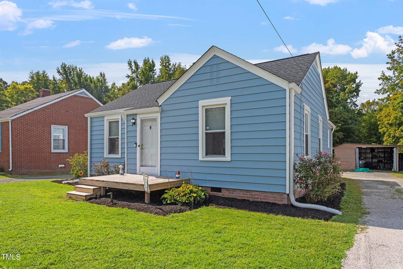 A small blue house with a gable roof, white trim, and a front porch. There are two flower beds with plants on either side of the porch. The house has a gravel driveway and neighboring houses are visible. The sky is clear and sunny.