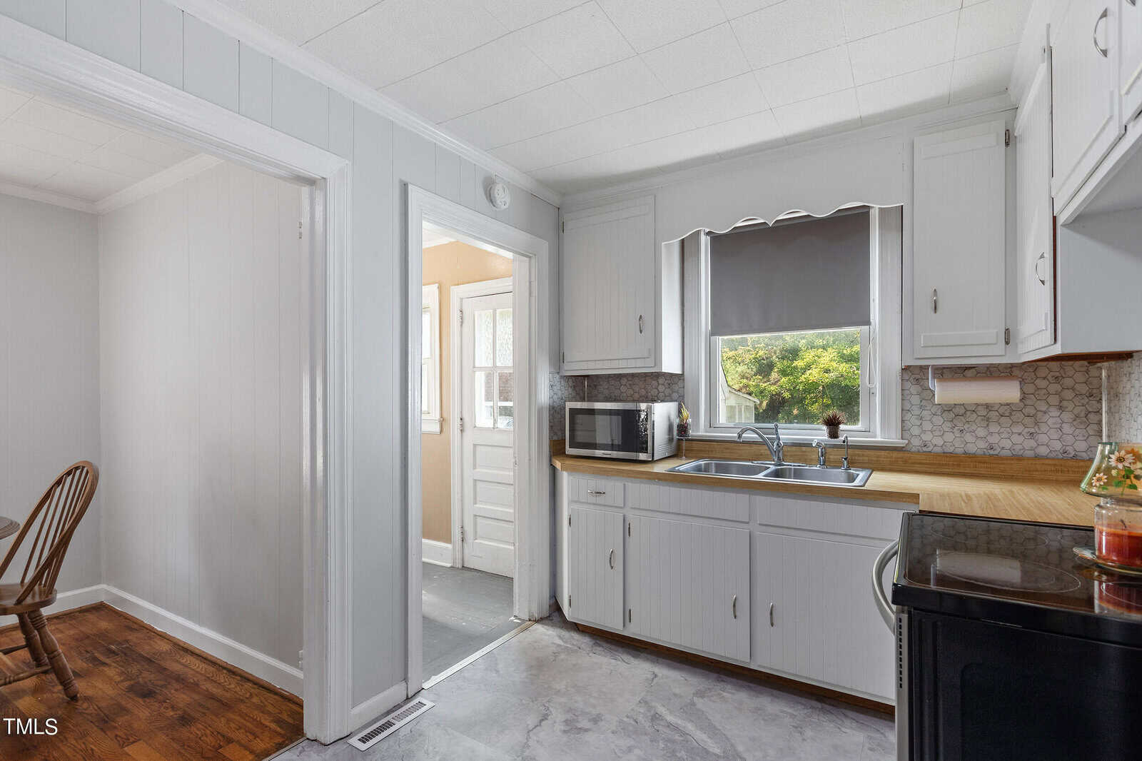 A bright kitchen with white cabinets, wooden countertops, and a double sink under a window with a gray shade. A microwave sits to the left, and a door leads outside. Hardwood flooring is visible in the adjoining room.