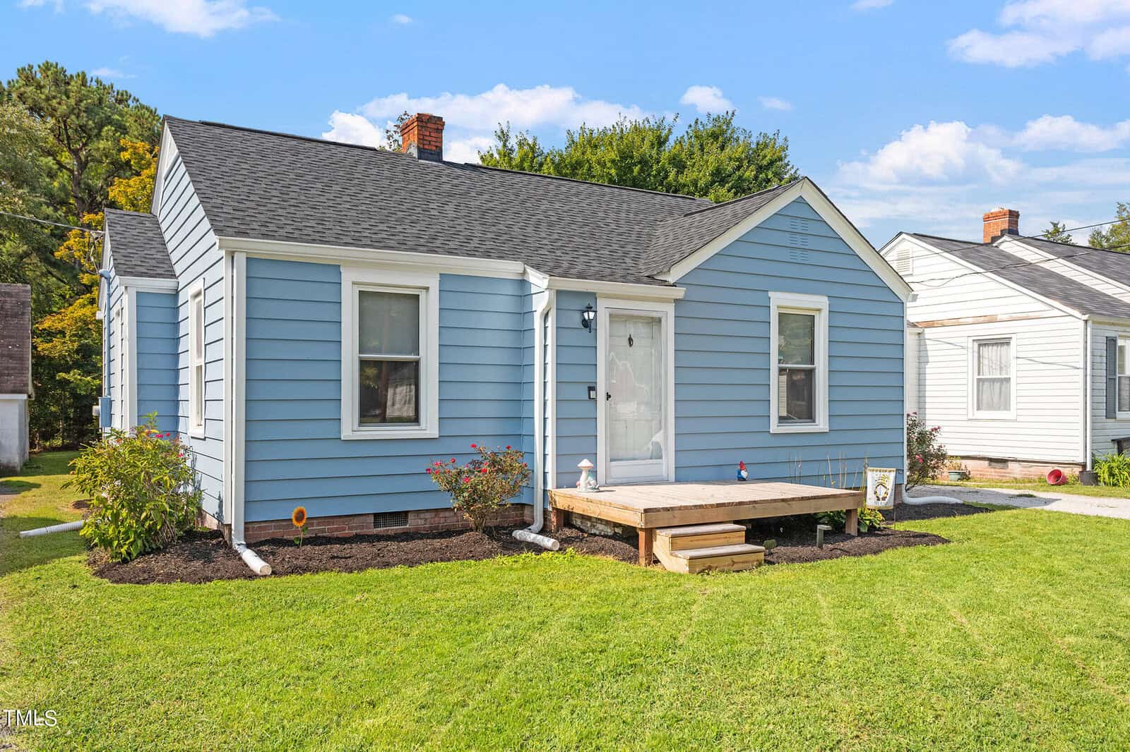 A small, charming light blue house with white trim and a black roof. It features a simple wooden front step, surrounded by a neatly maintained lawn and a few shrubs. The sky is clear and blue, enhancing the sunny feel of the scene.