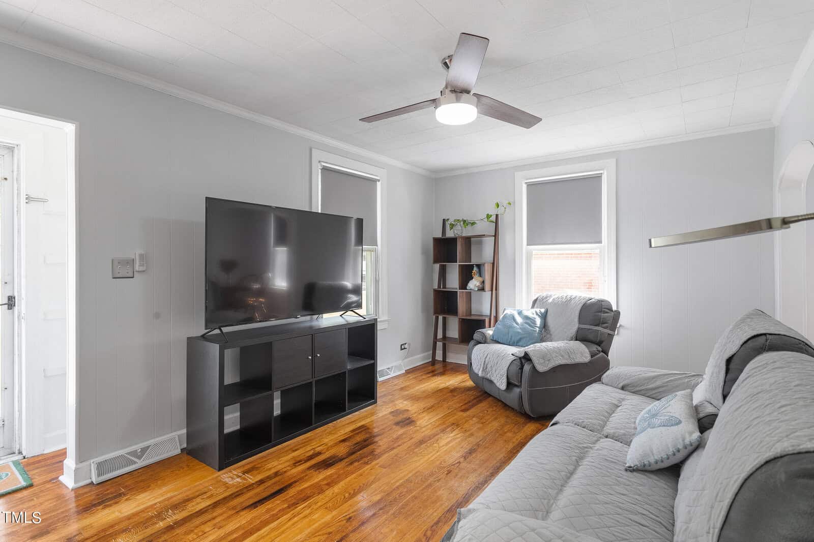 A cozy living room with a large flat-screen TV on a black stand. There's a gray loveseat and a matching reclining chair with pillows. The room has wooden floors, two windows with blinds, a ceiling fan, and a small bookshelf in the corner.