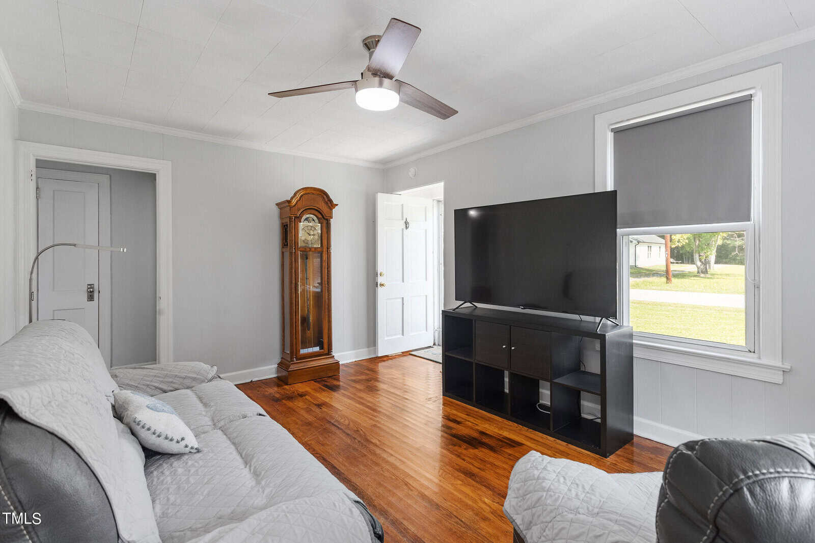 A living room with gray walls featuring a sofa and armchair, a large flat-screen TV on a black stand, and a wooden grandfather clock. A ceiling fan is on the white ceiling, and natural light comes through a window with a gray blind.