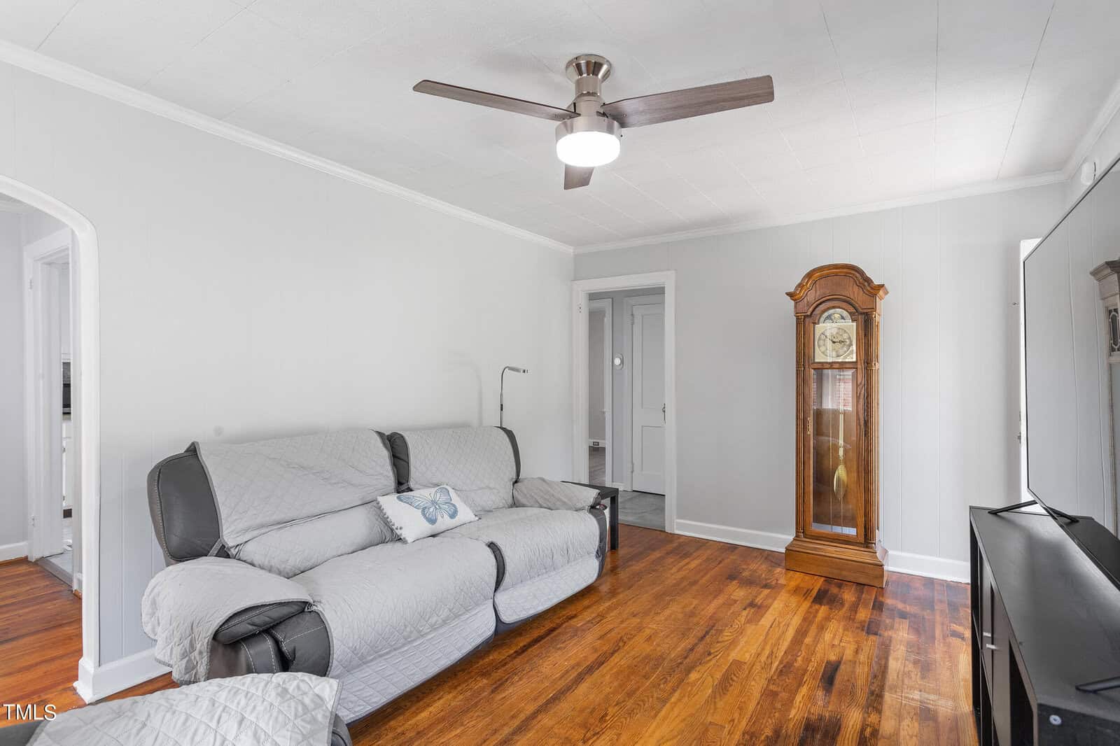 A cozy living room with a gray cushioned sofa, wooden floor, and a ceiling fan. A tall wooden grandfather clock stands near a doorway. Light gray walls and a white ceiling give the space a bright and airy feel.