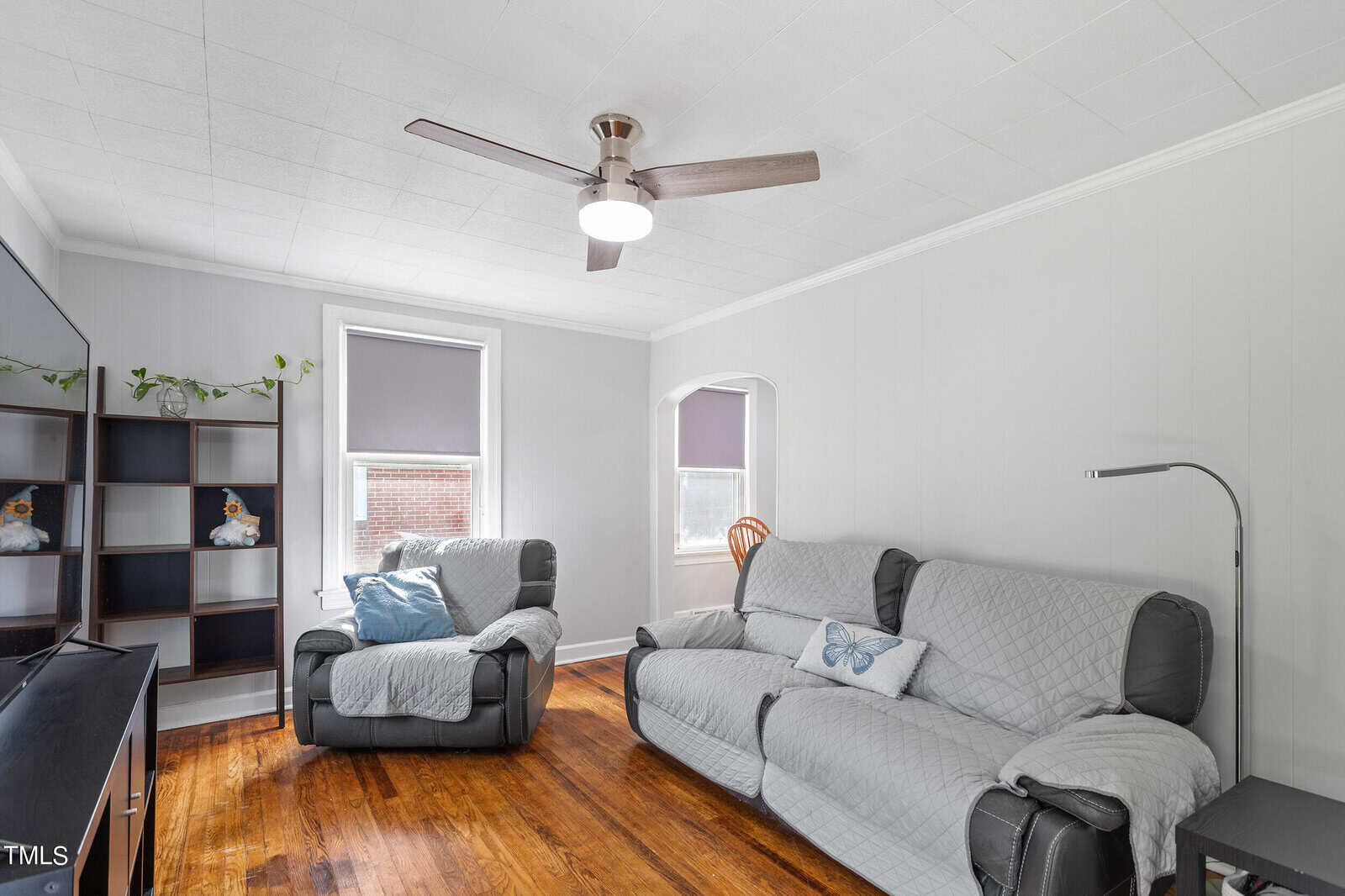 A cozy living room with gray walls features a gray sofa and armchair, both covered with quilted throws. A ceiling fan hangs above, and a shelf with plants and decorative items is by a window with closed blinds. A floor lamp stands nearby.