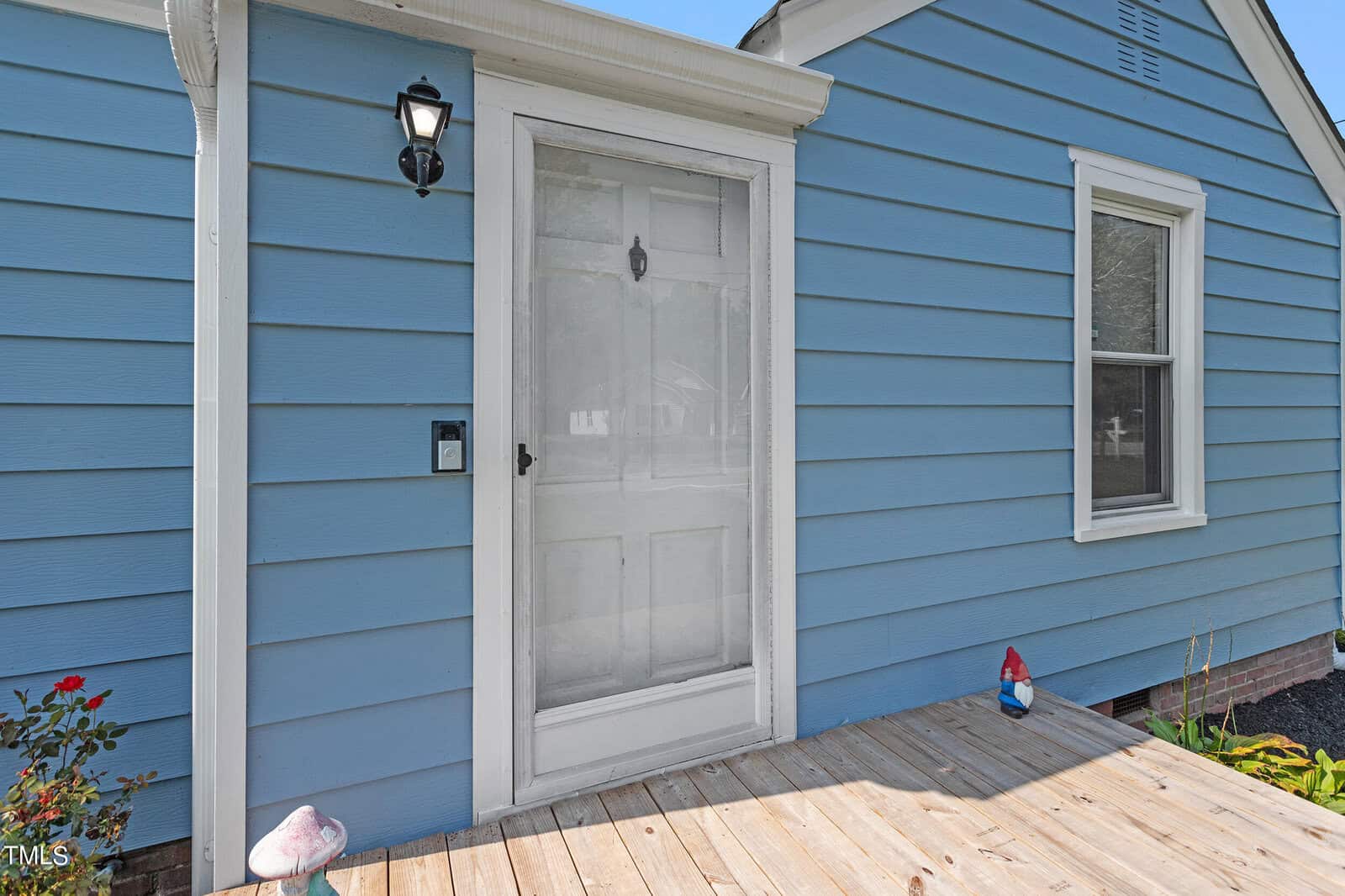 A small blue house with a wooden deck entrance, featuring a white door and screen door. There is a lantern-style light fixture beside the door and a decorative garden gnome on the deck. A window is visible to the right of the door.
