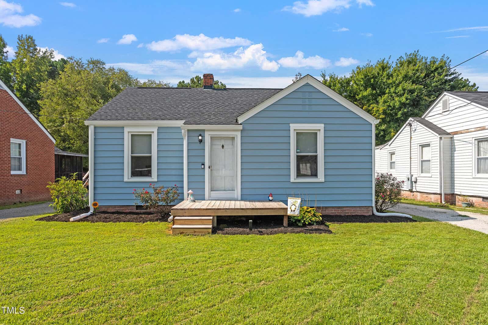 A small blue house with white trim sits on a neatly manicured lawn at 310 E Church Street, Creedmoor. It features a simple wooden porch and a white door. The sky is clear with a few clouds, and neighboring houses are partially visible.