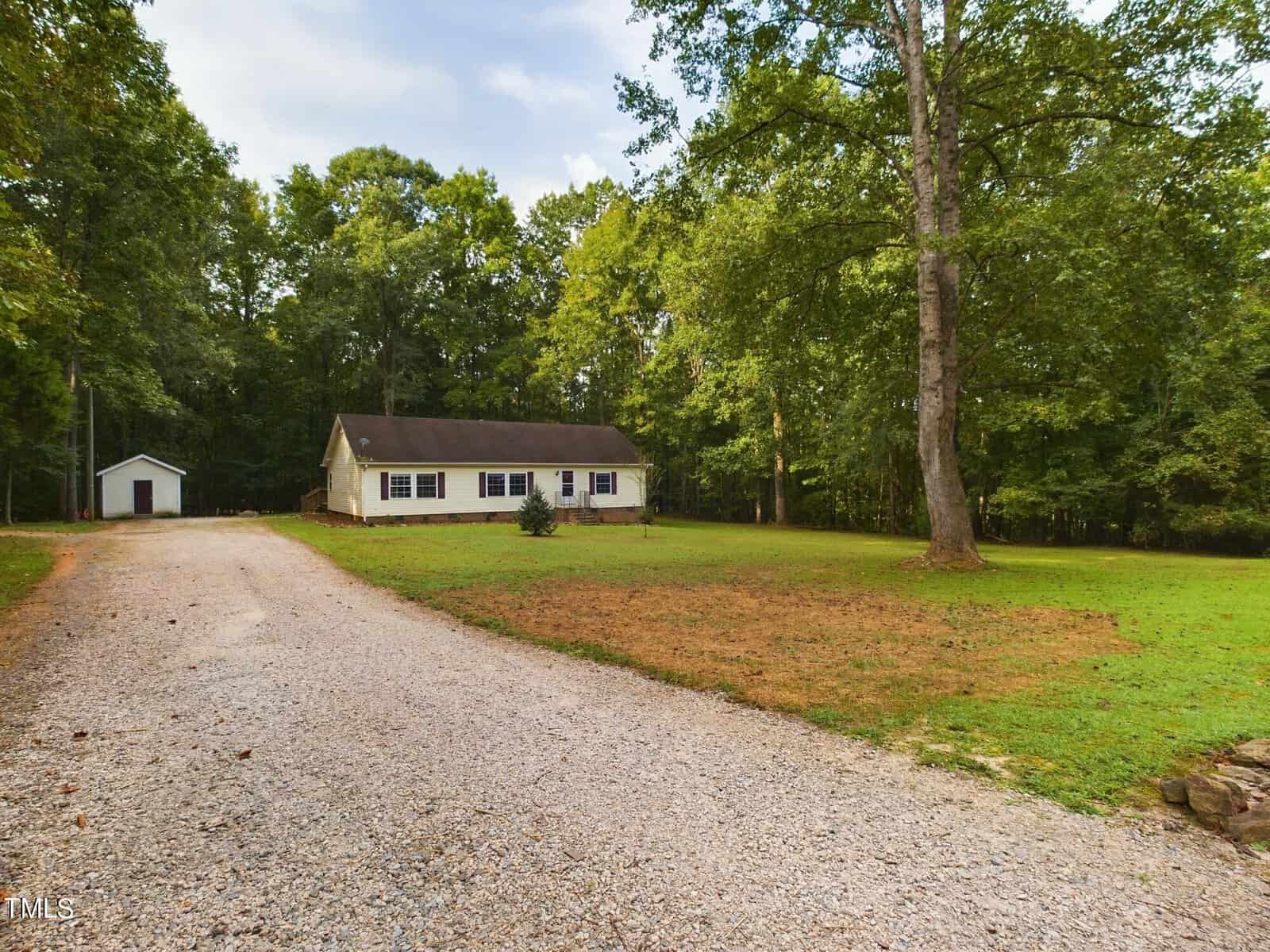 A single-story white house with a gabled roof is set in a green, wooded area. It is surrounded by a grassy lawn, and a gravel driveway leads up to the property. A small outbuilding is visible in the background, nestled among the trees.