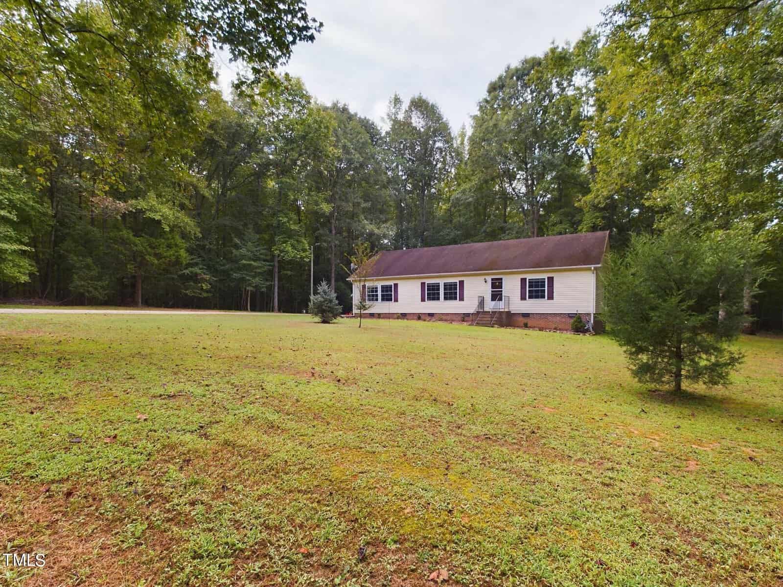 A single-story house with a light exterior and a dark roof is surrounded by a grassy lawn and trees. A small pathway leads to the front entrance. The area is wooded, providing ample greenery and privacy.