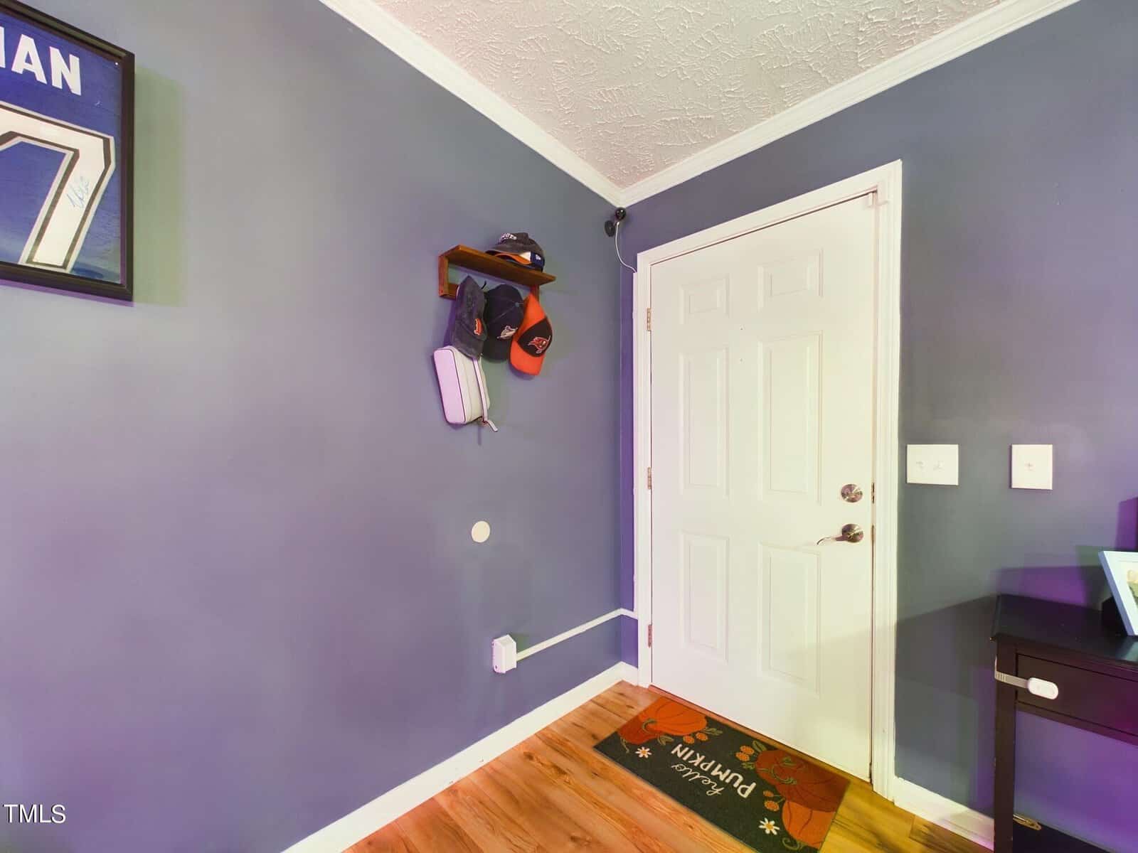 A corner of a room features a white door with a silver handle. A small shelf above the door holds hats and a bag. The dark blue walls contrast with a wooden floor, and a colorful doormat is placed at the entrance.