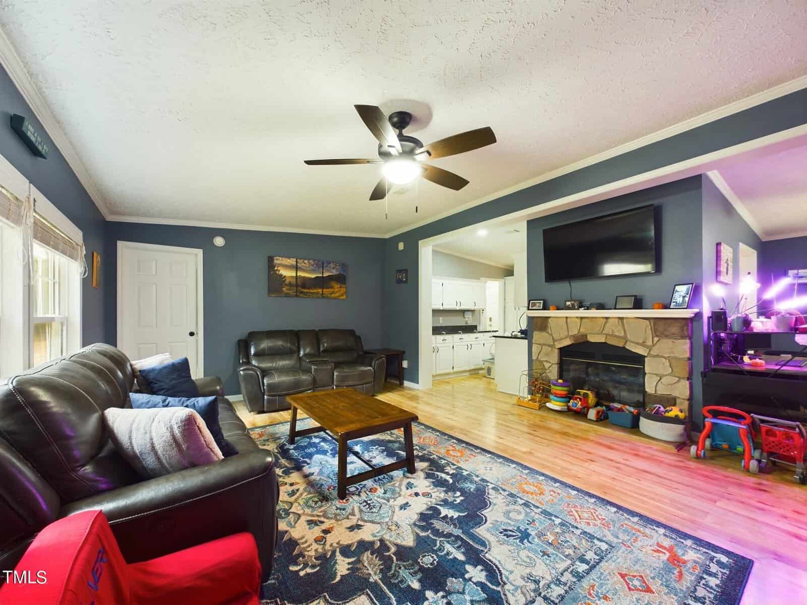 A cozy living room with dark blue walls, a ceiling fan, and hardwood floors. It features leather couches, a stone fireplace with a mounted TV, and a colorful rug. A kitchen is visible through an open doorway and children's toys are on the right.
