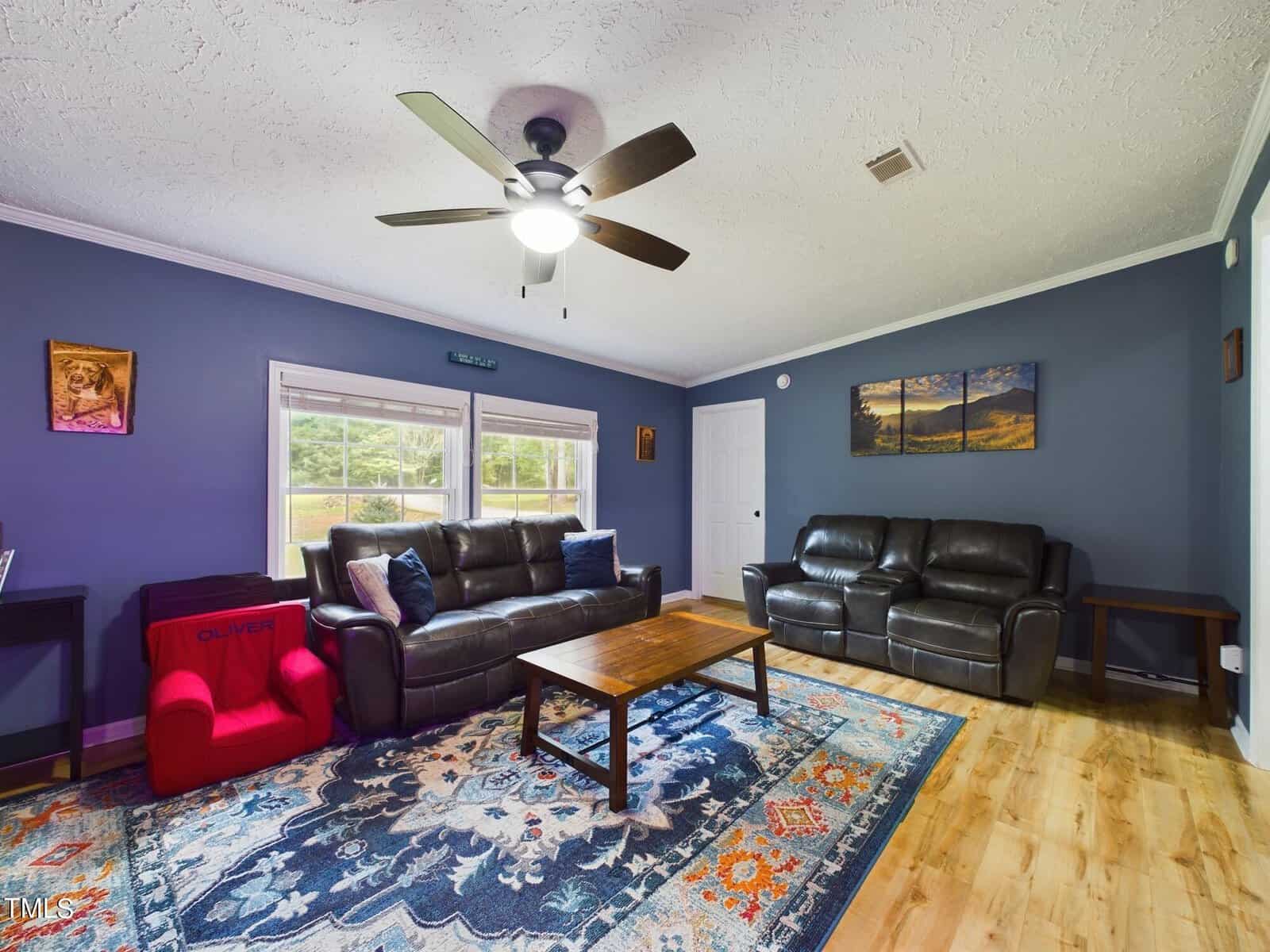 A living room with dark blue walls, hardwood floors, and ceiling fan. It features a black leather sofa and loveseat, a wooden coffee table, and a red children's chair. A colorful rug covers part of the floor. Three landscape pictures hang on the wall.