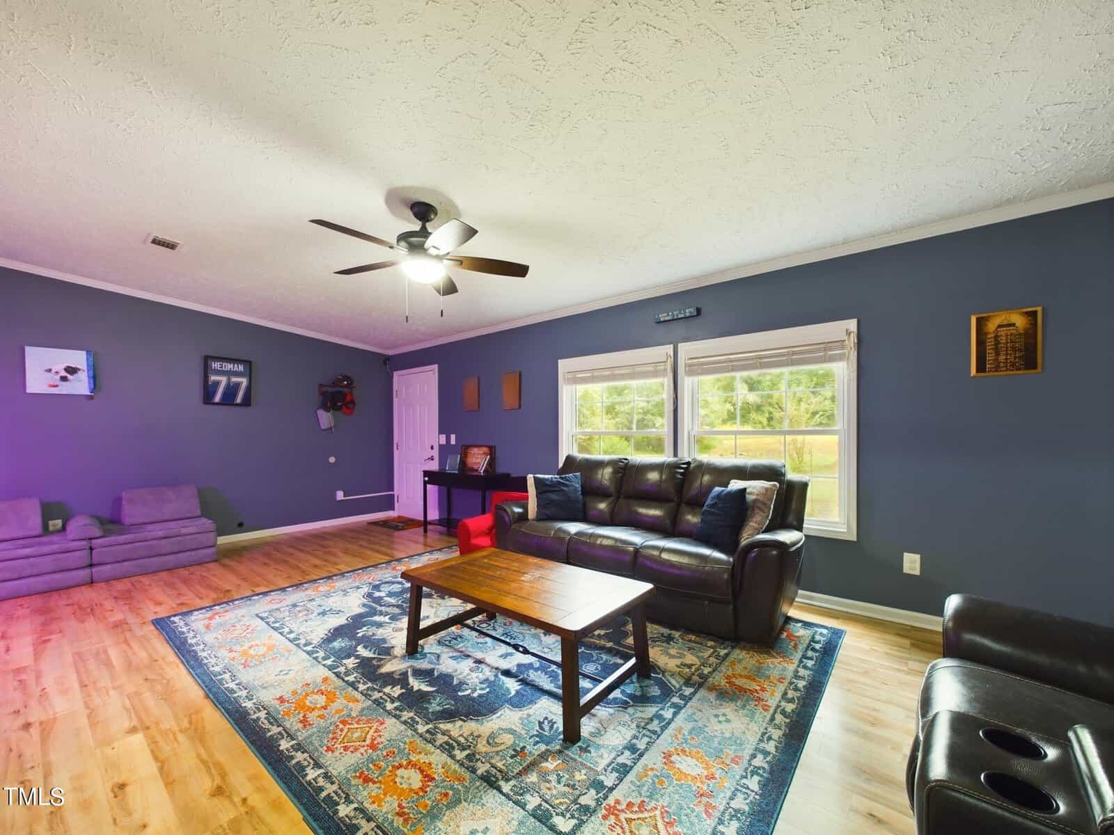 A living room with blue walls and light wood flooring features a brown leather sofa, a recliner, and a wooden coffee table on a colorful area rug. A ceiling fan is overhead, and framed decor is on the walls. Large windows let in natural light.