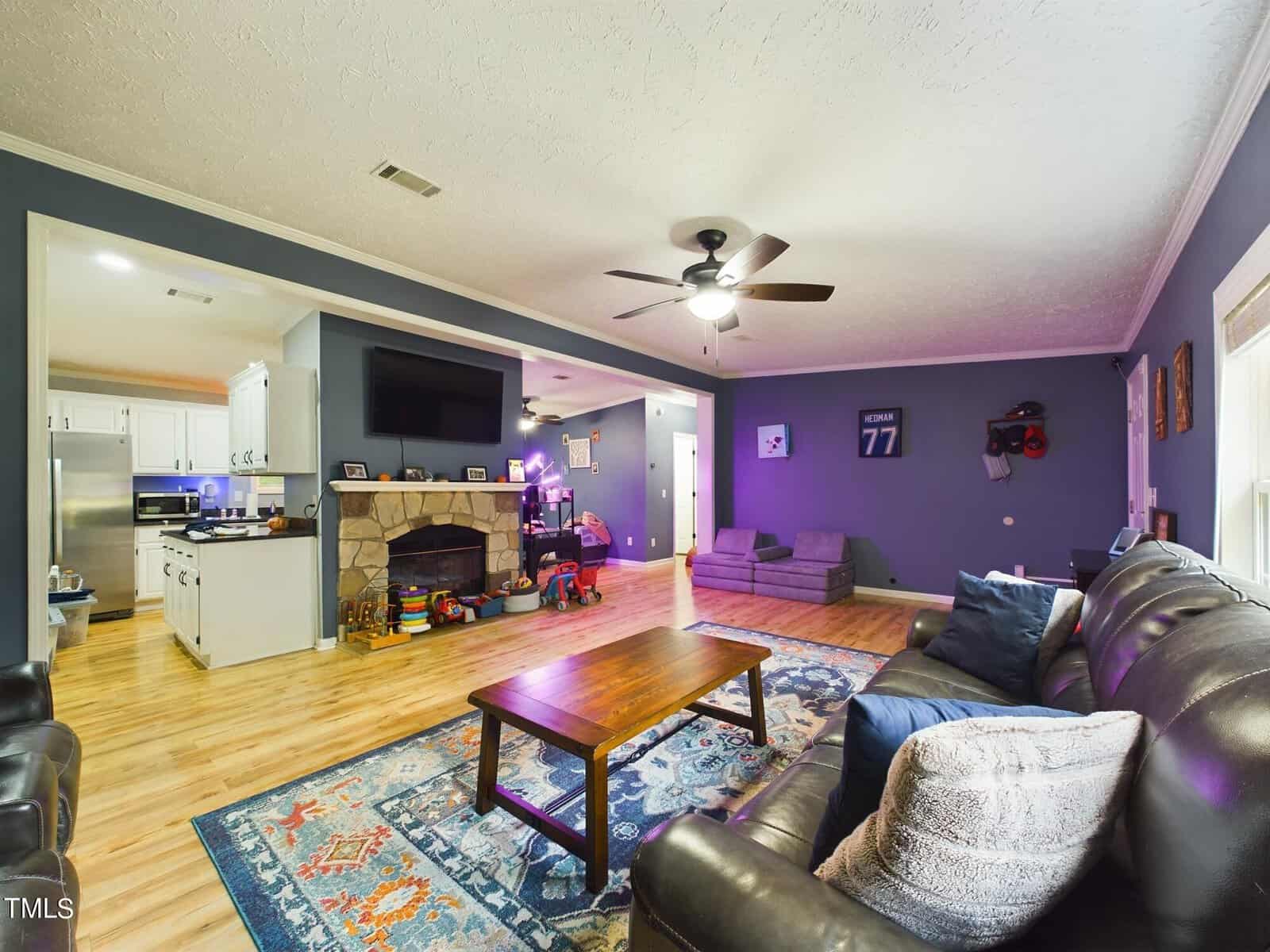 A spacious living room with hardwood floors and blue-gray walls. It features a leather sofa, wooden coffee table, and a large stone fireplace. The room has a ceiling fan and a flat-screen TV above the fireplace. The kitchen area is visible in the background.