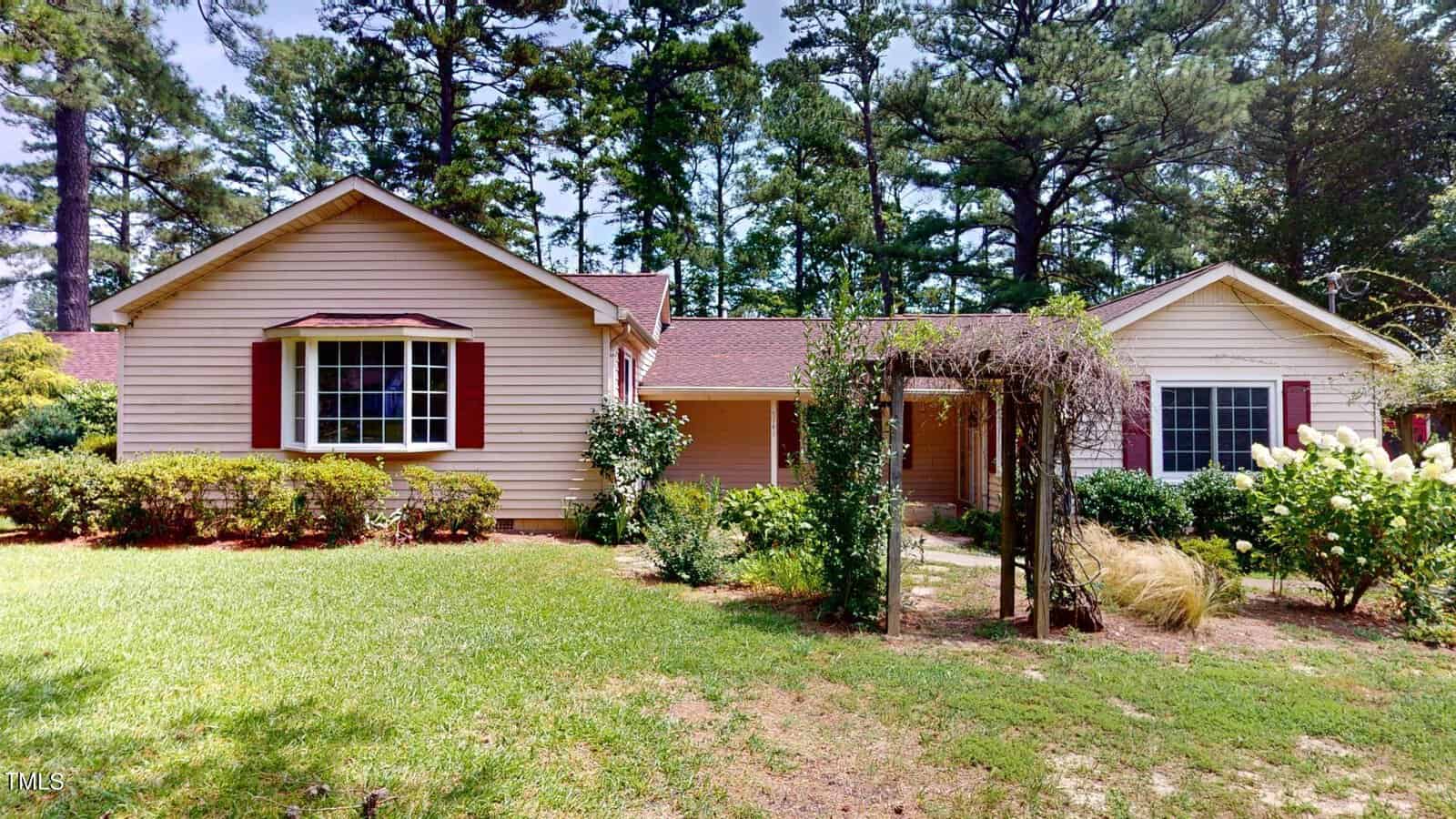 One-story tan house with a red roof and brown shutters, surrounded by bushes and trees. An arbor with climbing plants stands in front of the entrance, and the lawn is green and well-maintained.