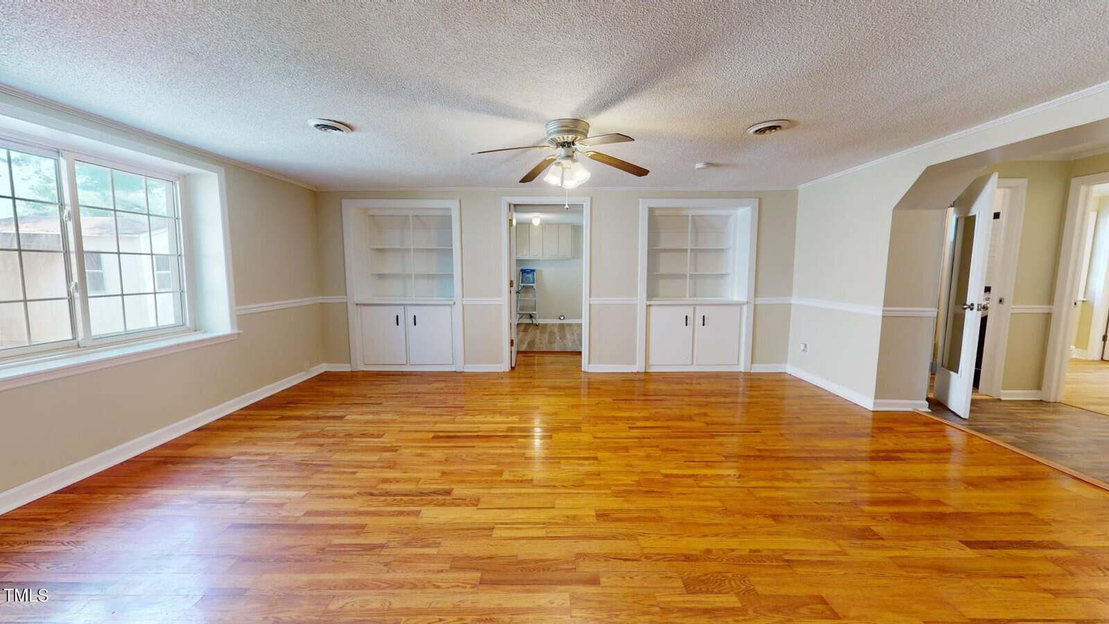 A spacious room with hardwood floors, large window on the left, ceiling fan with light, built-in shelves and cabinets on the far wall, and an open doorway leading to another room.