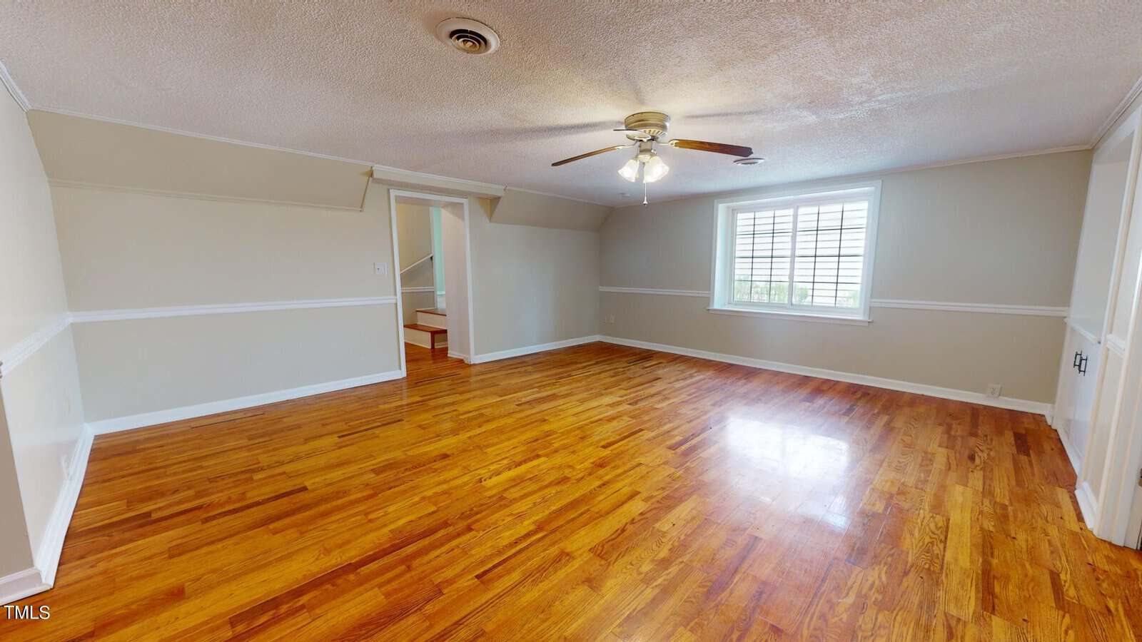Empty room with polished wooden floors, beige walls with white trim, a ceiling fan with lights, a window letting in natural light, and a small staircase leading up to another area.