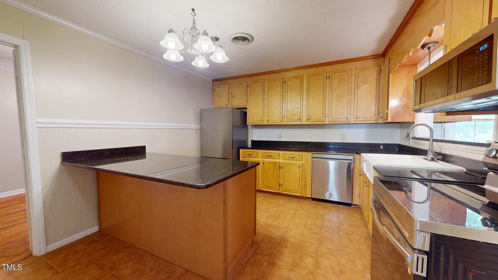 A kitchen with wood cabinets, black countertops, stainless steel appliances, a double sink, an island, tan tile flooring, and a light fixture hanging from the ceiling. A doorway leads to another room.