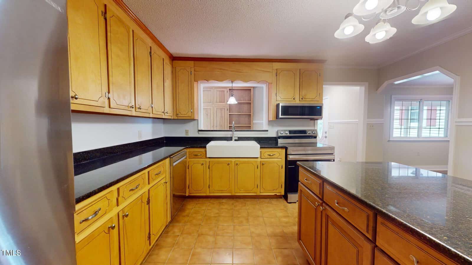 A kitchen with wooden cabinets, black countertops, a farmhouse sink, stainless steel appliances, tile flooring, and a central island under ceiling lights. An arched doorway leads to another room.