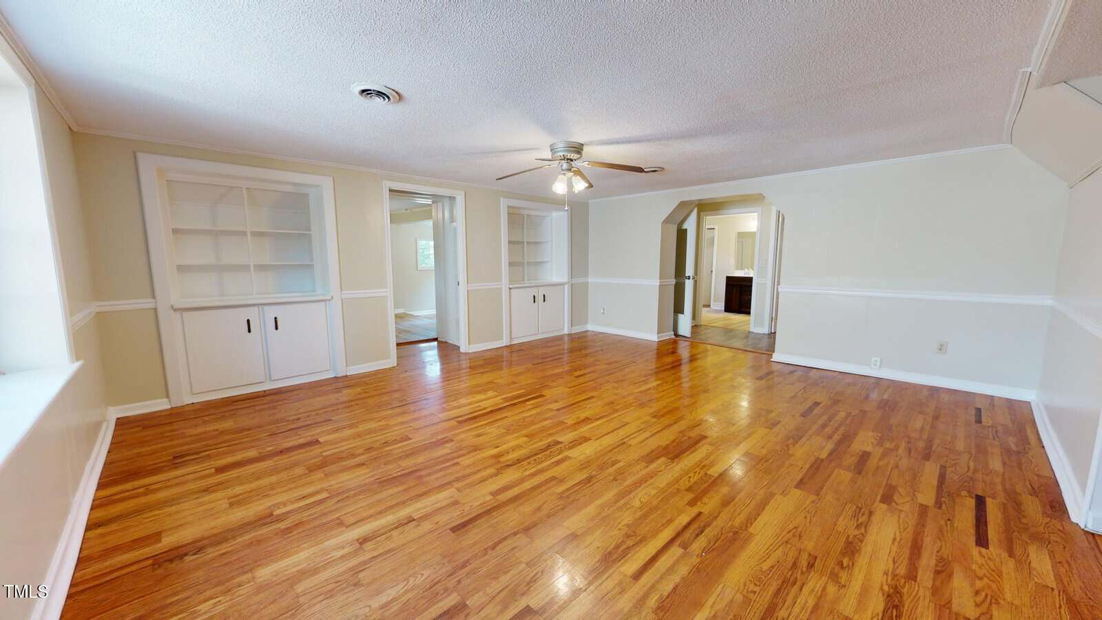 Empty living room with hardwood floors, white walls, built-in shelves with cabinets, a ceiling fan with lights, and large windows letting in natural light. Archway leads to another room.