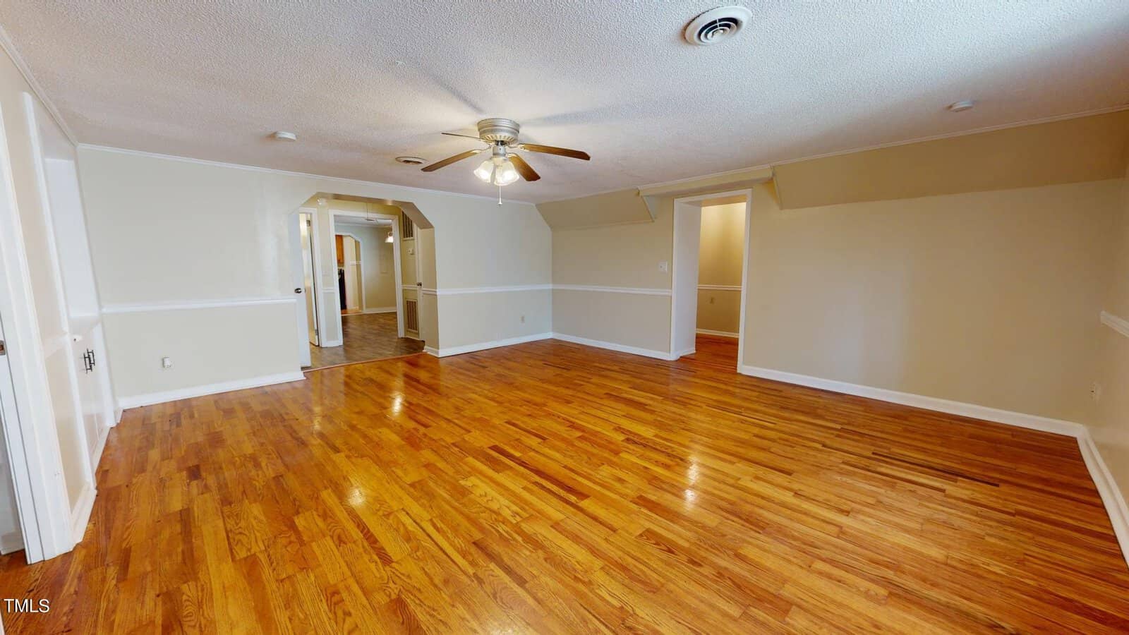 Empty room with polished hardwood floors, beige walls with white trim, a ceiling fan with lights, two doorways, and a window allowing natural light. The ceiling is partially sloped near the walls.