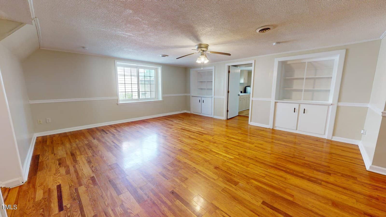 A spacious room with polished hardwood floors, neutral walls, a ceiling fan with lights, a large window, and built-in white shelves with cabinets on one side. An open doorway leads to another area.