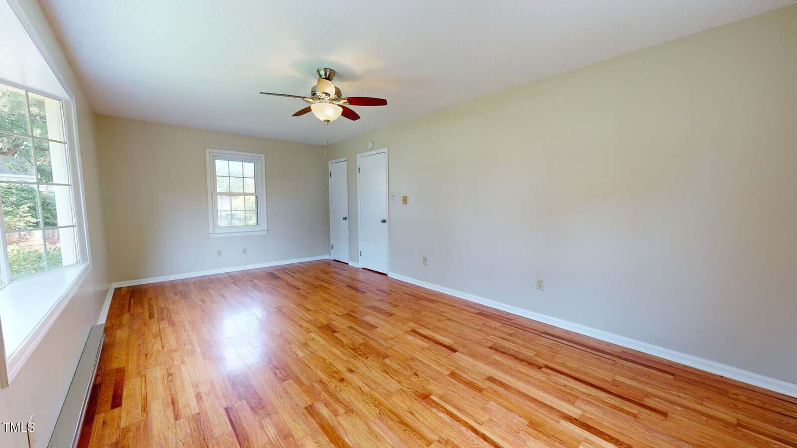 An empty room with light beige walls, wooden floors, a ceiling fan with a light, two windows on one wall, and two white doors on the opposite wall. Natural light comes in through the windows.