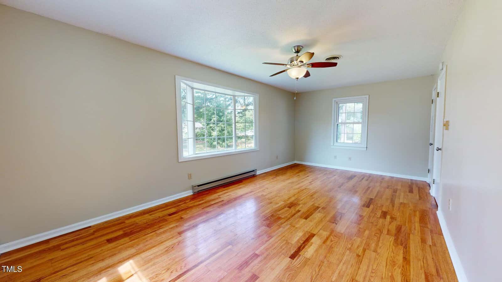 A spacious, empty room with light beige walls, a large window, a smaller window, wooden floors, a white ceiling, a ceiling fan with lights, and a closed door on the right wall.