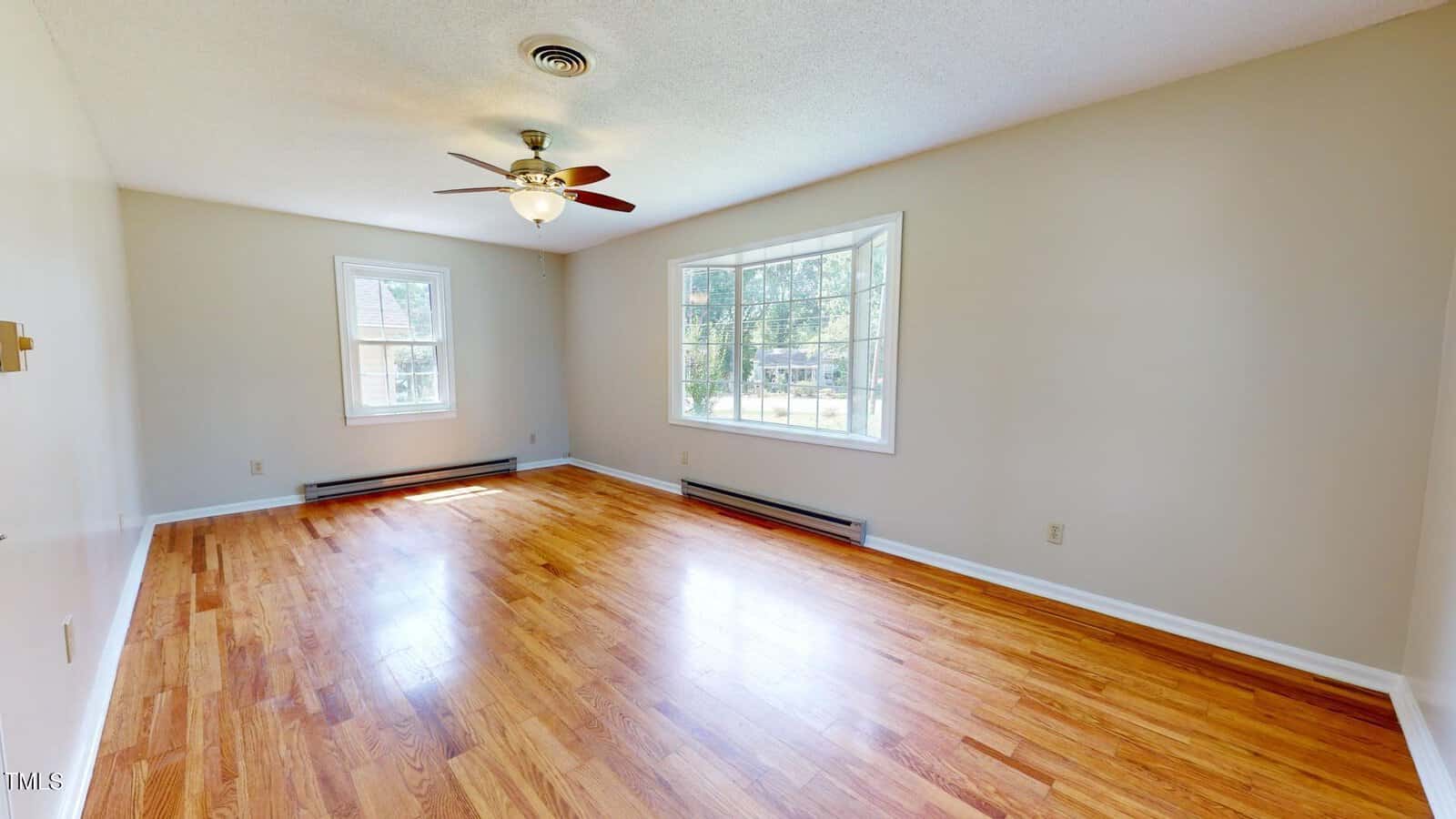 Empty room with hardwood floors, beige walls, large window, ceiling fan with light, and baseboard heaters beneath the windows. Natural light fills the space.
