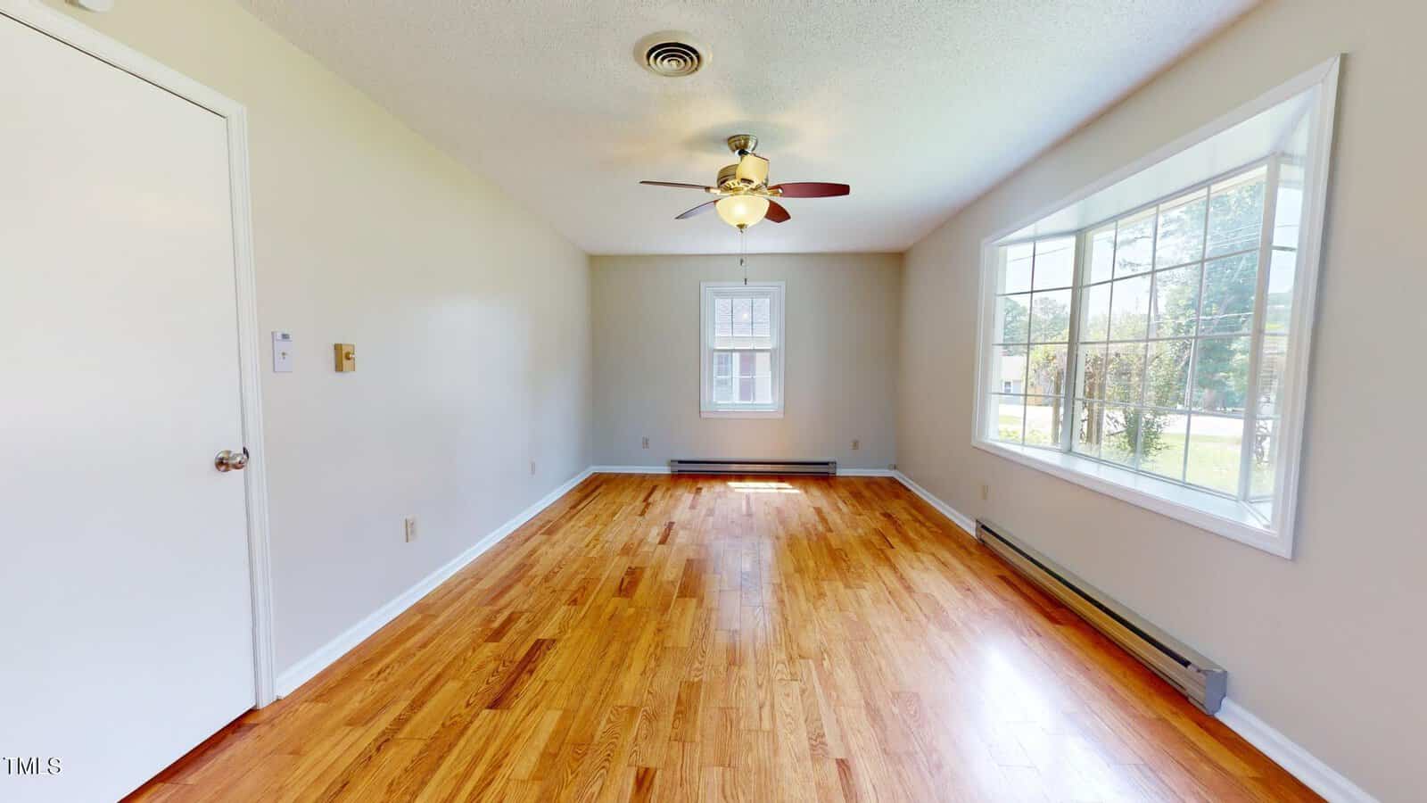 Bright, empty room with hardwood floors, beige walls, ceiling fan, large window on the right letting in sunlight, and a smaller window at the far end.